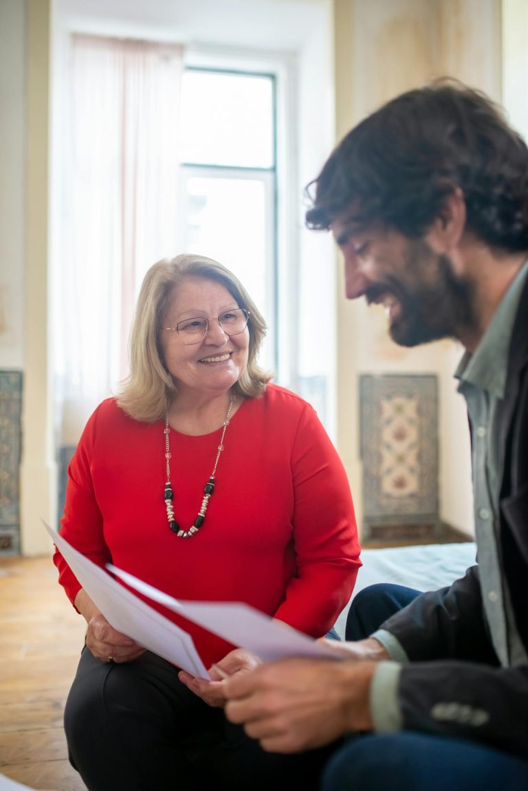 Elderly woman and bearded man discussing documents in a sunlit room.